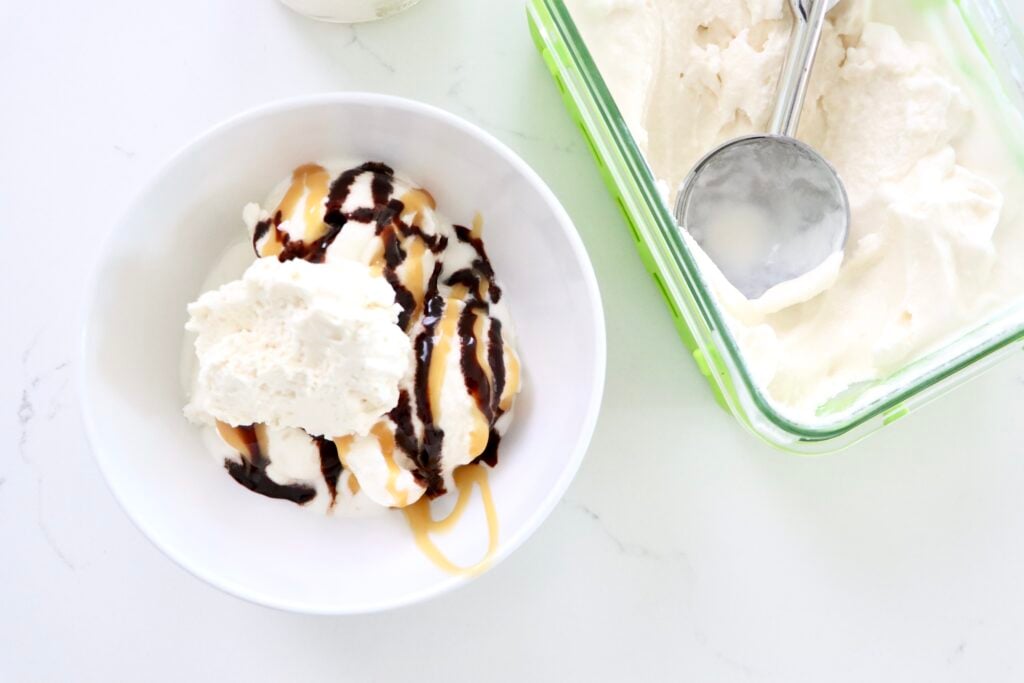 Homemade Vanilla Ice cream in a bowl with chocolate and caramel sauce topped with whipped cream next to a container of vanilla ice cream