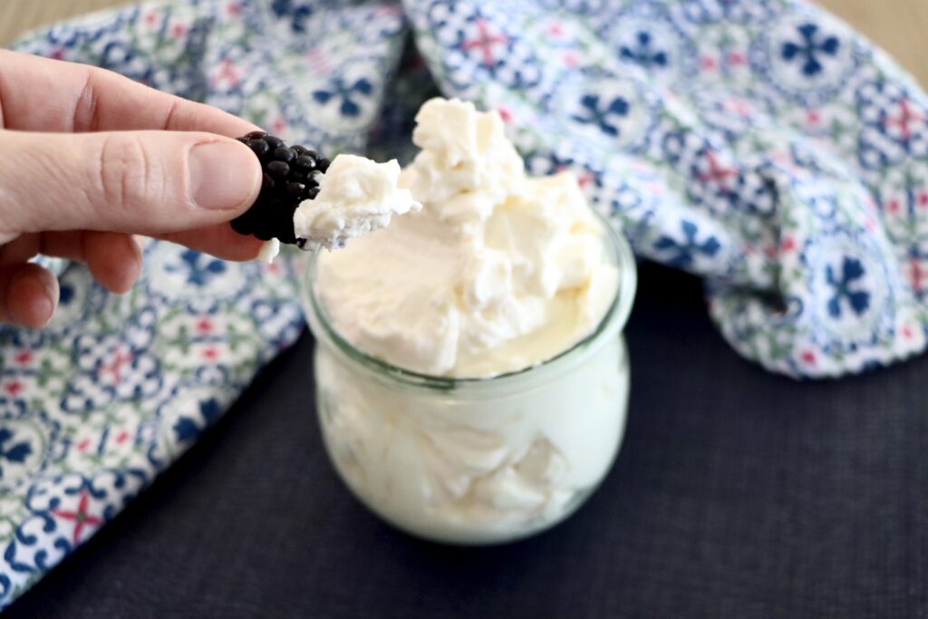 Stabilized Whipped Cream in a glass jar and a hand holding a blackberry dipped in whipped cream