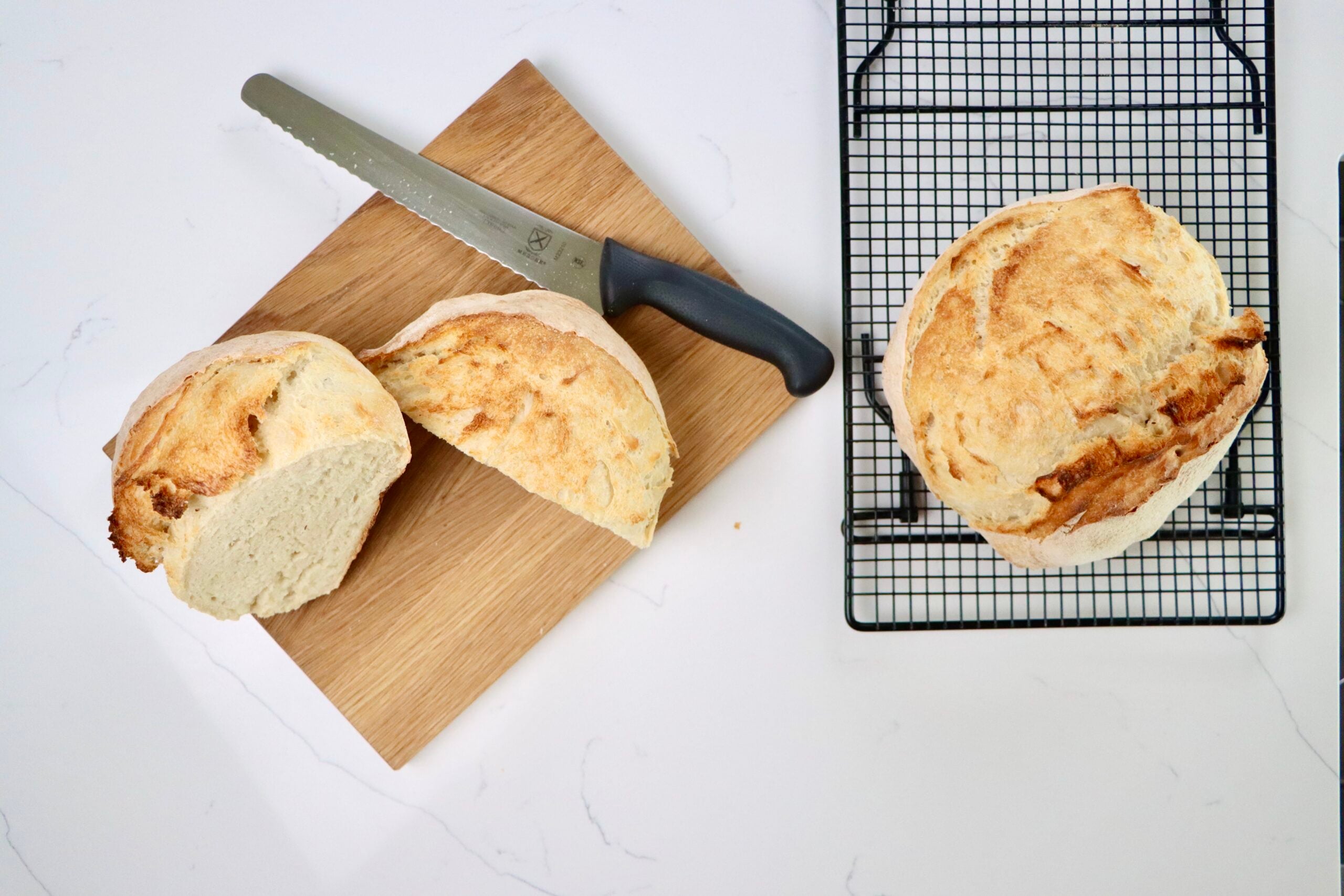 a loaf of sourdough bread cut in half on a cutting board with a knife and a loaf of sourdough bread on a cooling rack