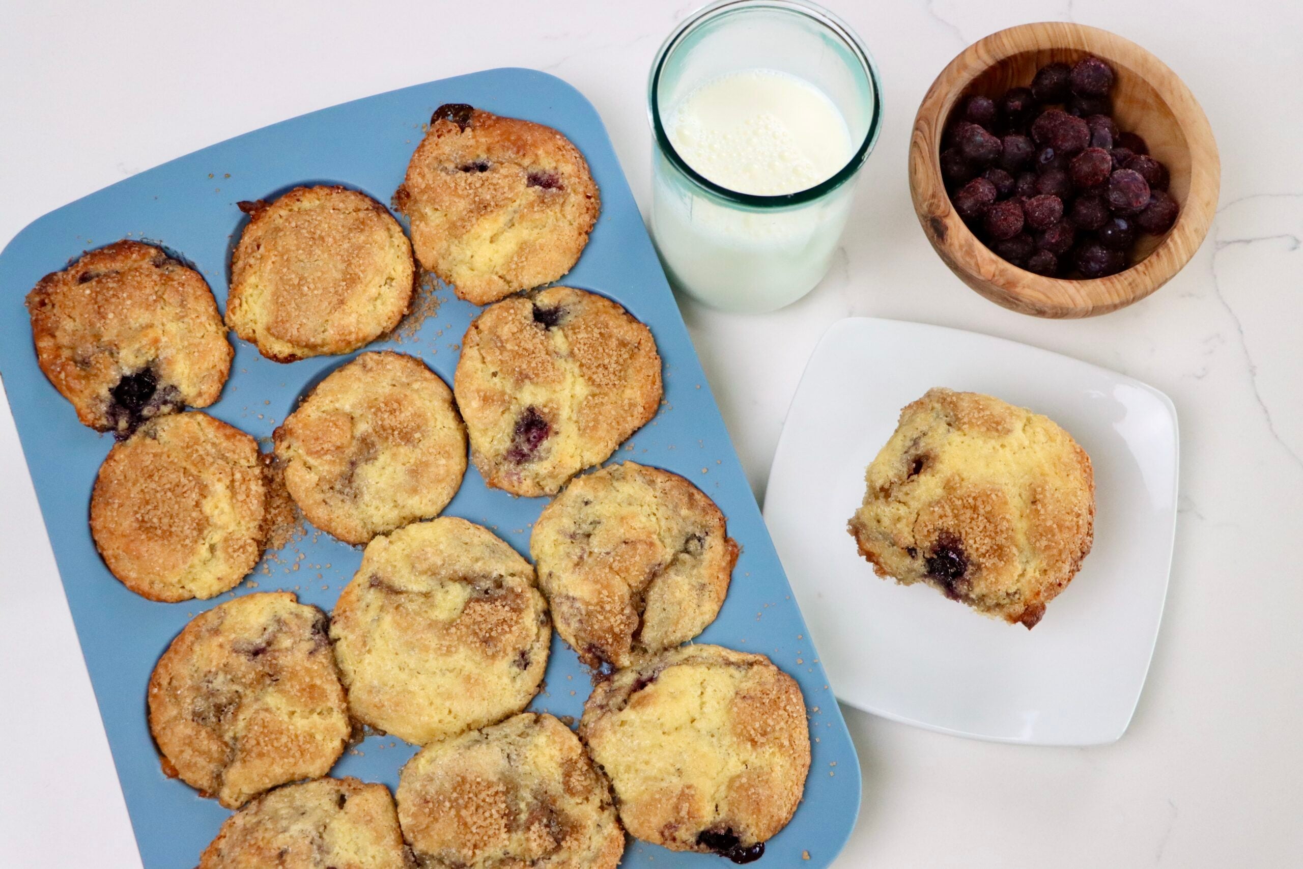 Sourdough Einkorn Blueberry Muffins in a muffin tin with a bowl of blueberries, a single muffin on a plate and a glass of milk