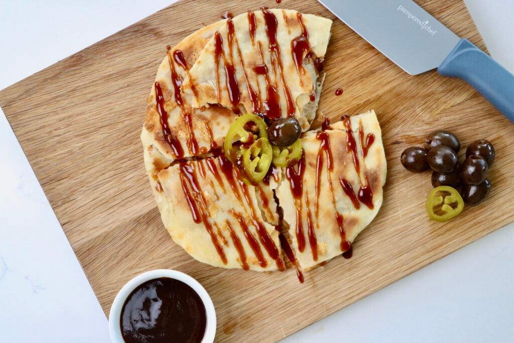 BBQ Chicken Quesadilla on a cutting board with a knife and a bowl of BBQ sauce, garnished with jalepenos and olives