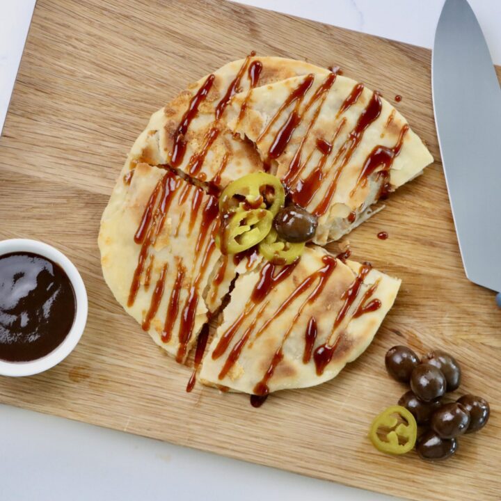 BBQ Chicken Quesadilla on a cutting board with a knife and a bowl of BBQ sauce, garnished with jalepenos and olives