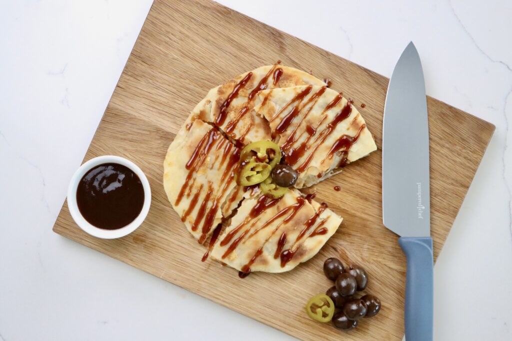 BBQ Chicken Quesadilla on a cutting board with a knife and a bowl of BBQ sauce, garnished with jalepenos and olives
