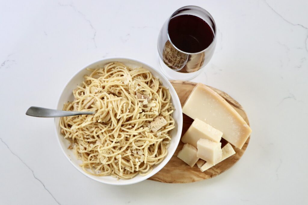 Chicken spaghetti in a bowl on a cutting board with parmesan cheese and a glass of wine