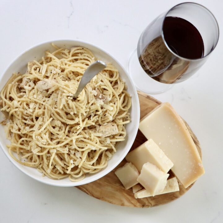 Chicken spaghetti in a bowl on a cutting board with parmesan cheese and a glass of wine
