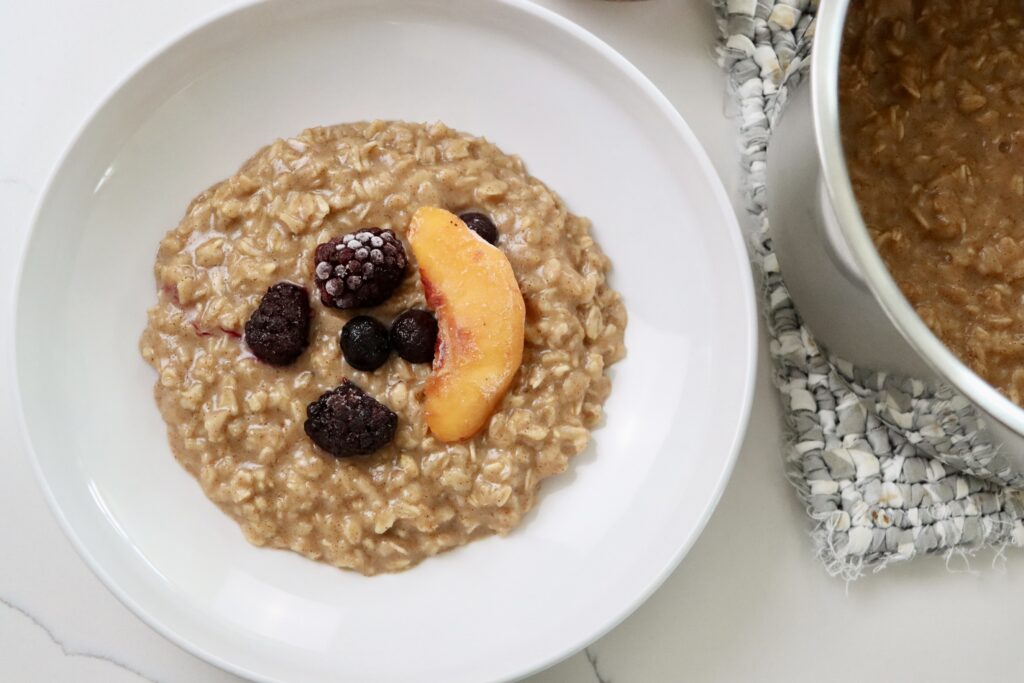 Maple Cinnamon Oatmeal in a bowl with fruit on top and a bowl of fruit and a pot of oatmeal on a hot pad