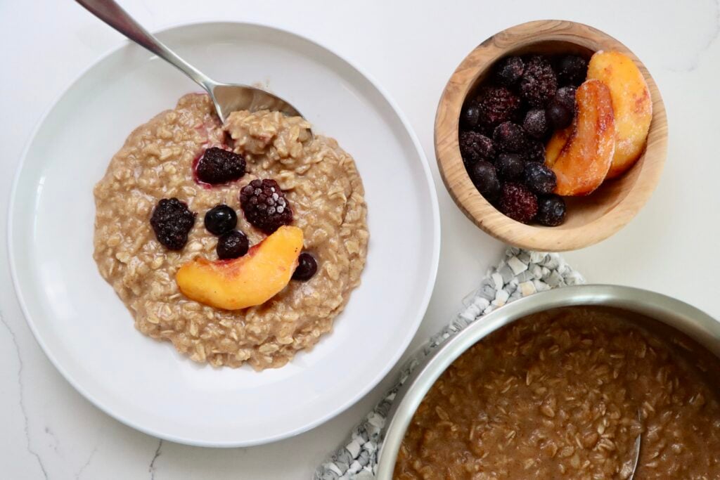 Maple Cinnamon Oatmeal in a bowl with fruit on top and a bowl of fruit and a pot of oatmeal on a hot pad
