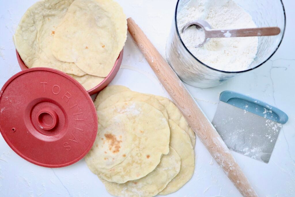 Sourdough Tortillas in a tortilla warmer next to a rolling pin, dough scraper and a jar of flour