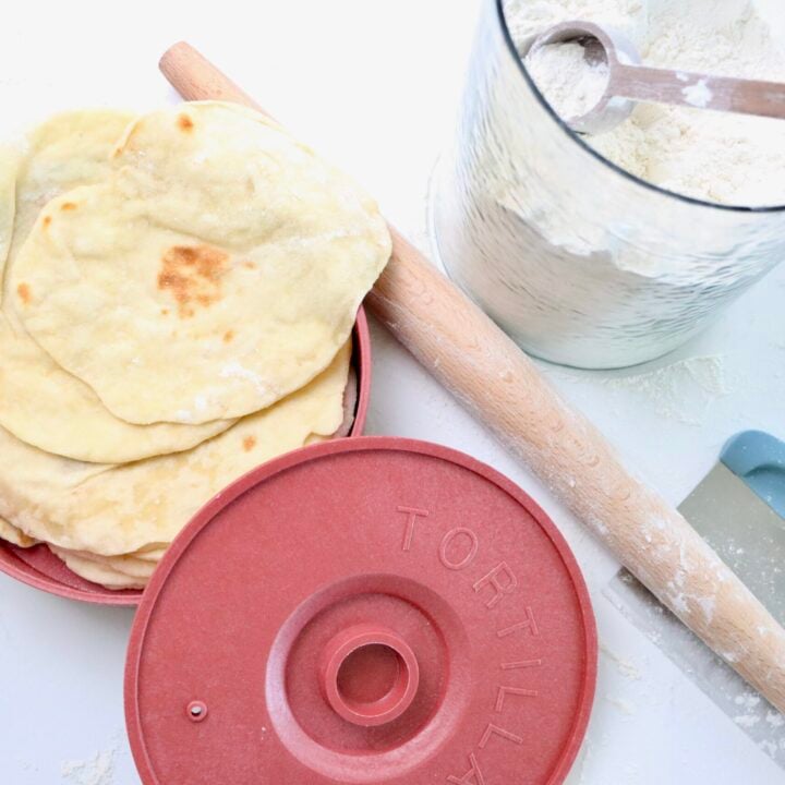 Sourdough Tortillas in a tortilla warmer next to a rolling pin, dough scraper and a jar of flour