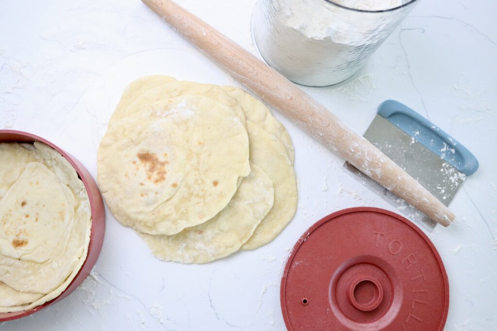 Sourdough Tortillas in a tortilla warmer next to a rolling pin, dough scraper and a jar of flour