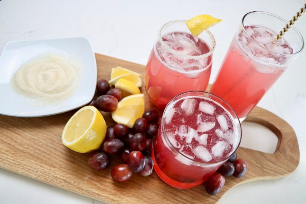 three cups of sparkling grape juice on a cutting board with lemons and grapes and a plate of sugar