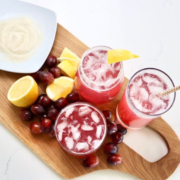 three cups of sparkling grape juice on a cutting board with lemons and grapes and a plate of sugar