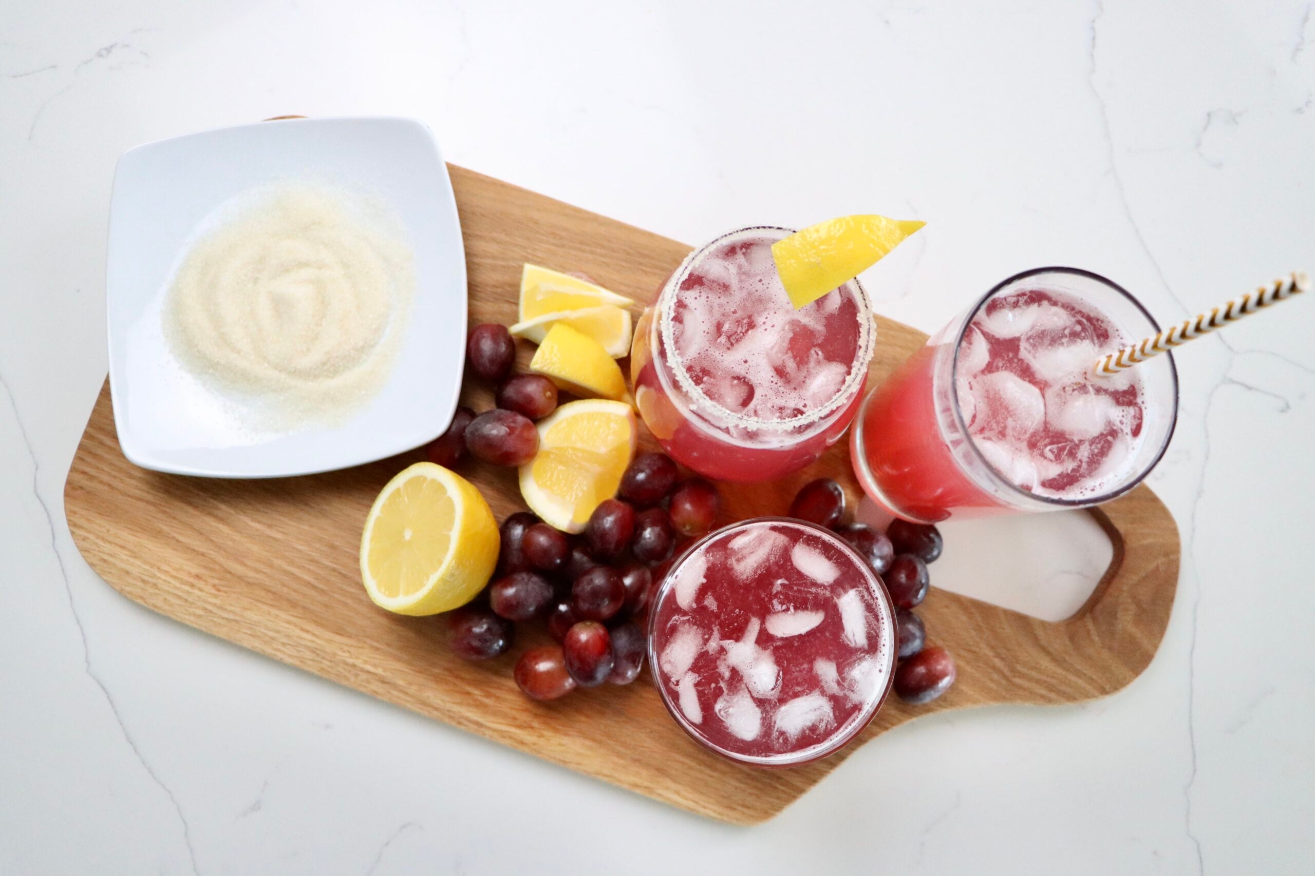 three cups of sparkling grape juice on a cutting board with lemons and grapes and a plate of sugar