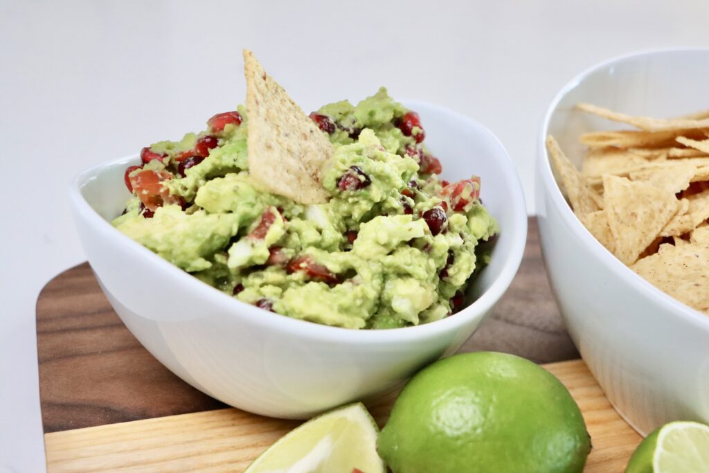 Pomegranate Guacamole in a bowl and chips in a bowl sitting on a cutting board garnished with tomatoes and limes