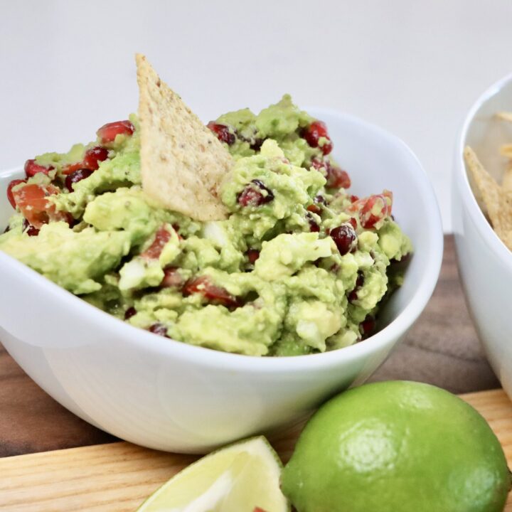 Pomegranate Guacamole in a bowl and chips in a bowl sitting on a cutting board garnished with tomatoes and limes