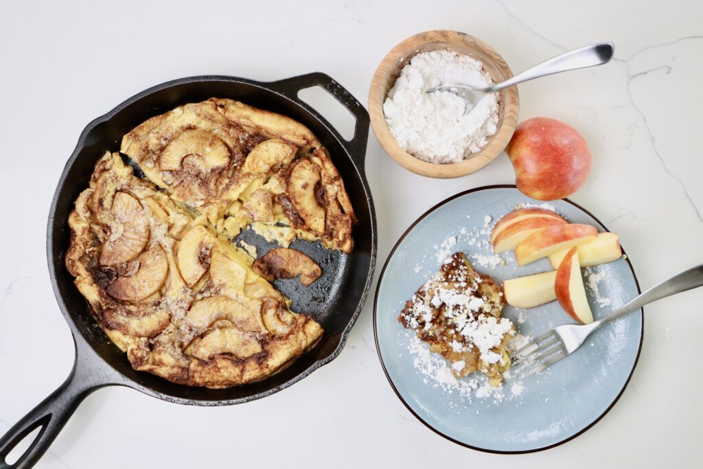 Sourdough Apple Dutch Baby in a skillet and on a plate with some sliced apples and a bowl of powdered sugar