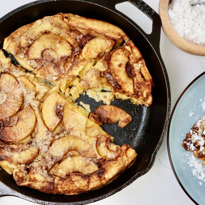 Sourdough Apple Dutch Baby in a skillet and on a plate with some sliced apples and a bowl of powdered sugar