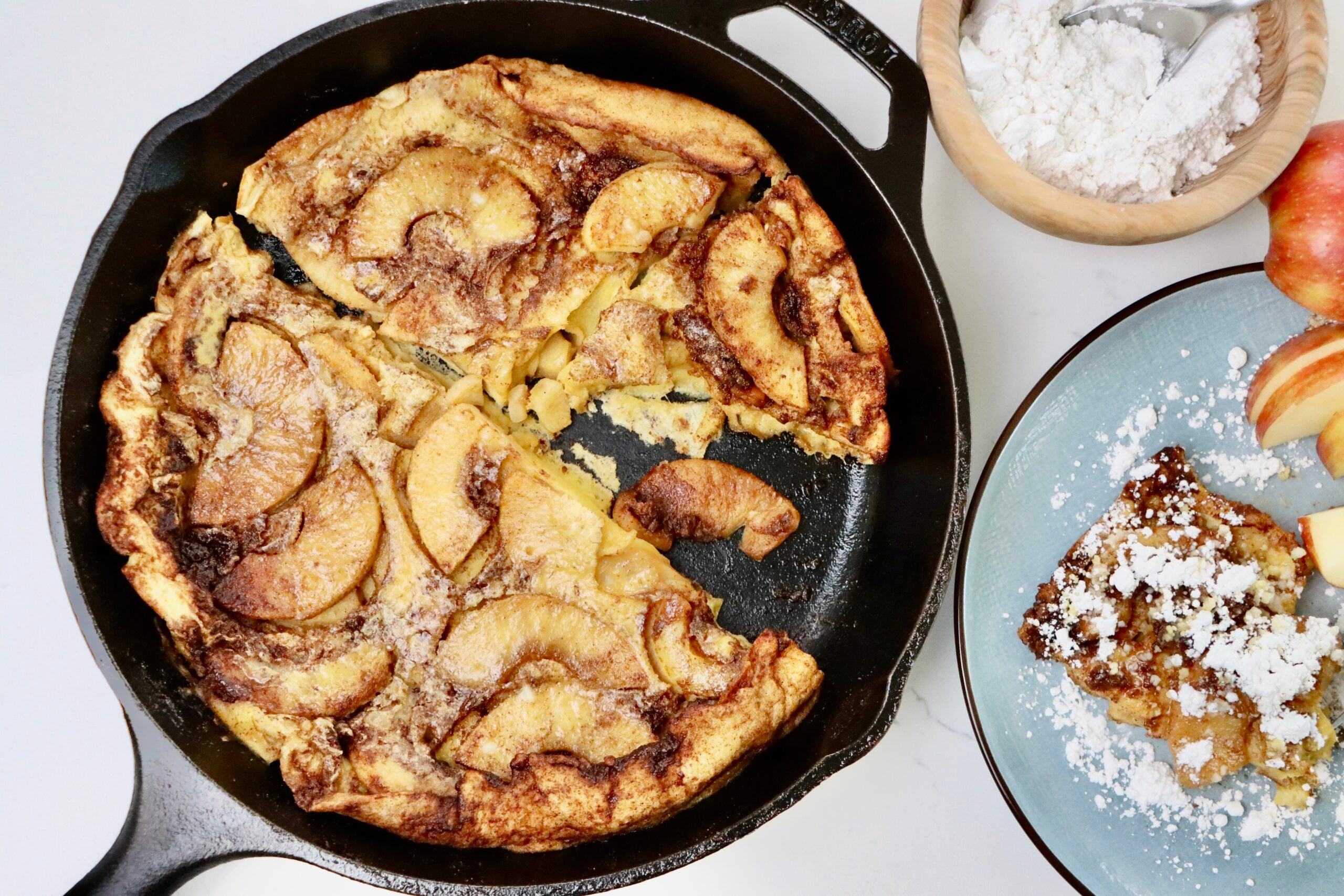 Sourdough Apple Dutch Baby in a skillet and on a plate with some sliced apples and a bowl of powdered sugar