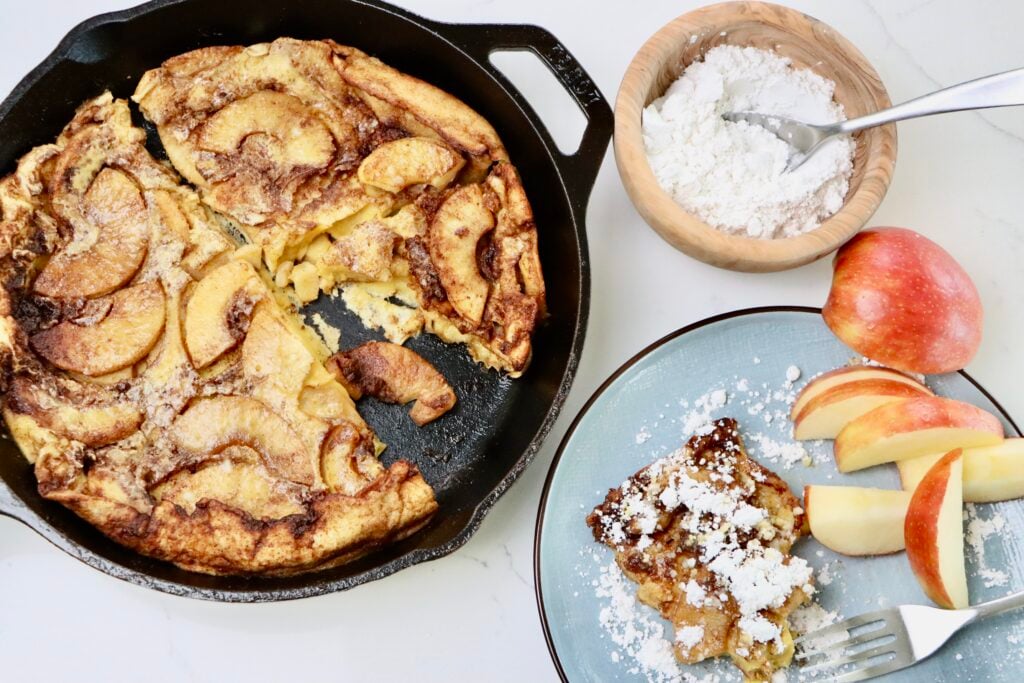 Sourdough Apple Dutch Baby in a skillet and on a plate with some sliced apples and a bowl of powdered sugar