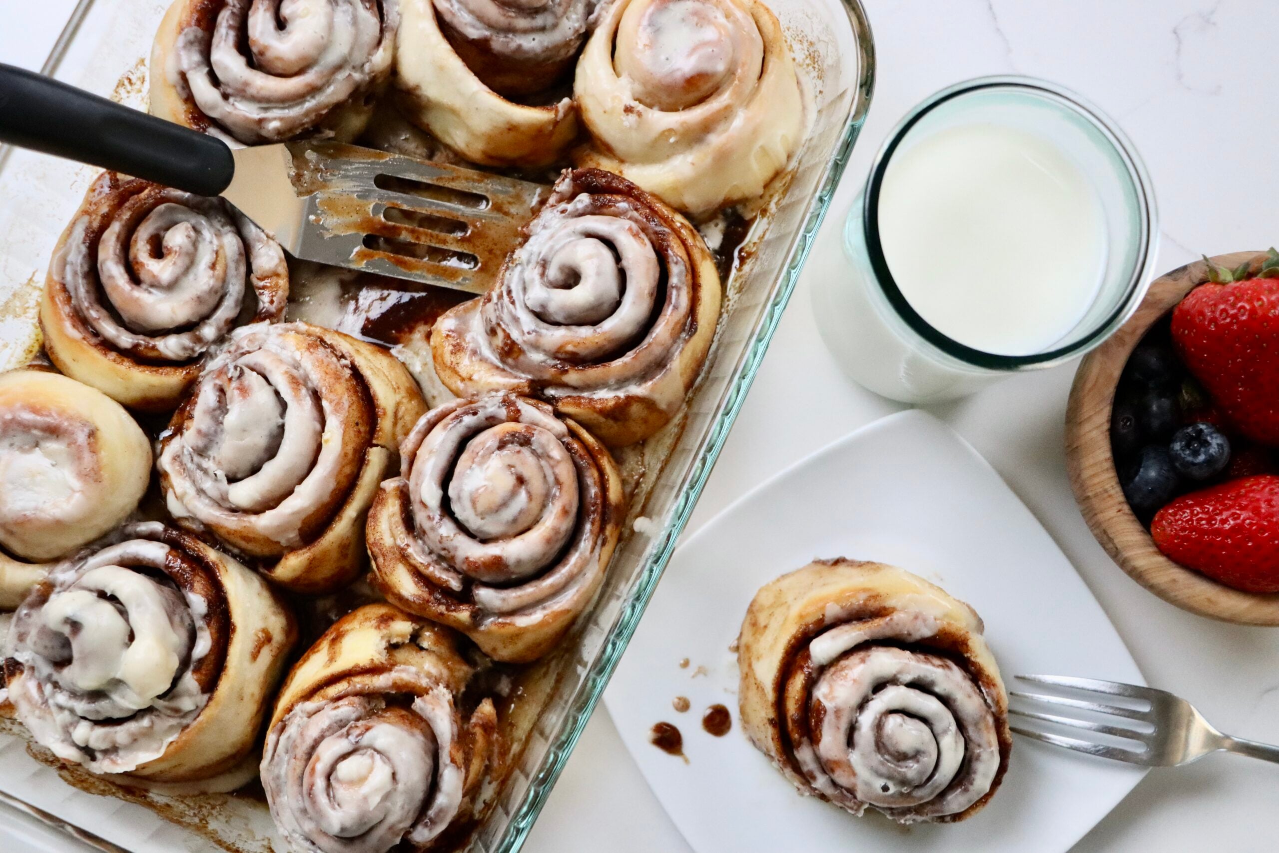 Sourdough Cinnamon Rolls in a pan with one cinnamon roll on a plate sitting next to a bowl of berries and a glass of milk.