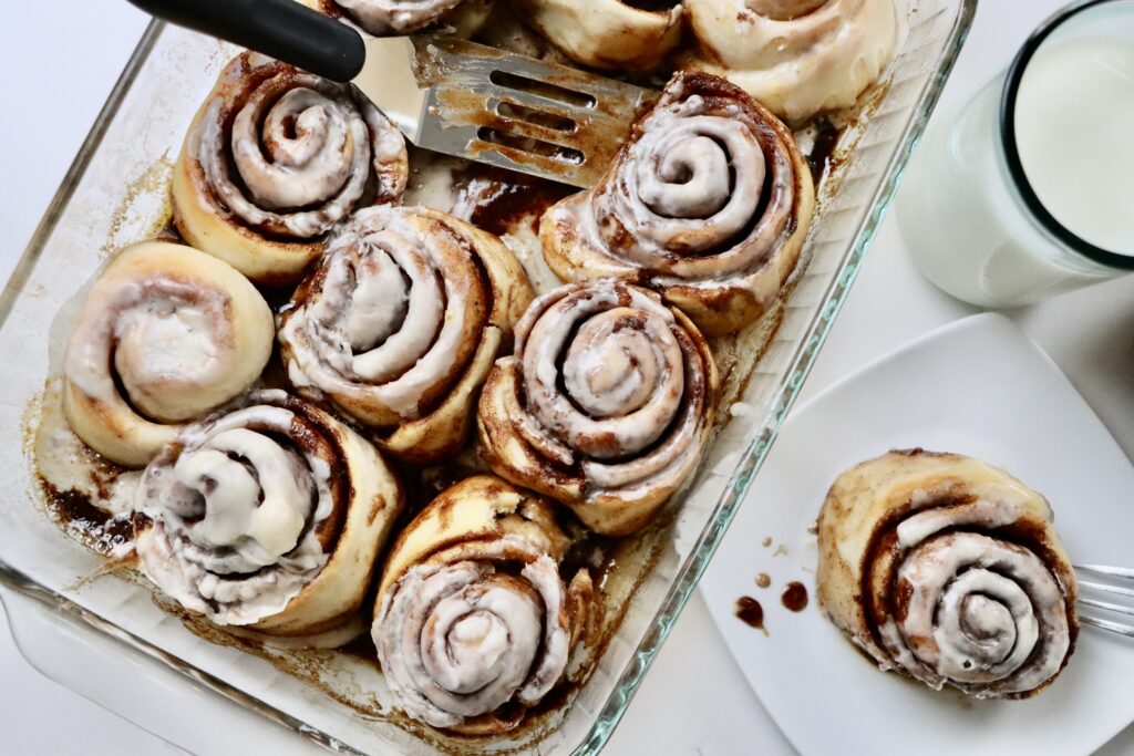 Sourdough Cinnamon Rolls in a pan with one cinnamon roll on a plate sitting next to a bowl of berries and a glass of milk.