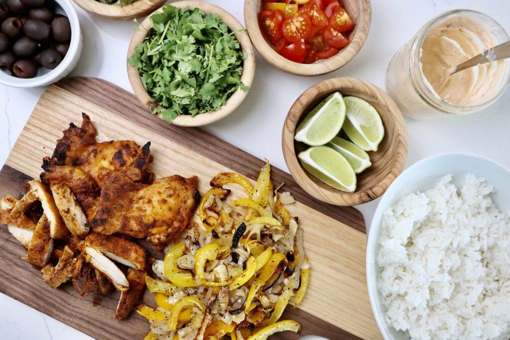 A bowl of homemade chicken fajita bowl with jasmine rice, roasted bell peppers, lettuce, tomatoes, and a dollop of chipotle crema, garnished with fresh cilantro. Surrounded by fresh toppings and ingredients on a white background.