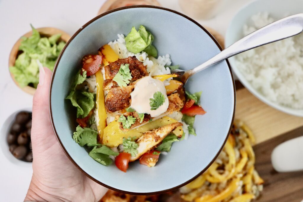 A bowl of homemade chicken fajita bowl with jasmine rice, roasted bell peppers, lettuce, tomatoes, and a dollop of chipotle crema, garnished with fresh cilantro. Surrounded by fresh toppings and ingredients on a white background.