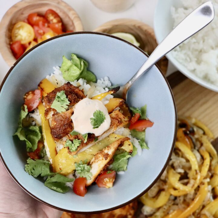 A bowl of homemade chicken fajita bowl with jasmine rice, roasted bell peppers, lettuce, tomatoes, and a dollop of chipotle crema, garnished with fresh cilantro. Surrounded by fresh toppings and ingredients on a white background.