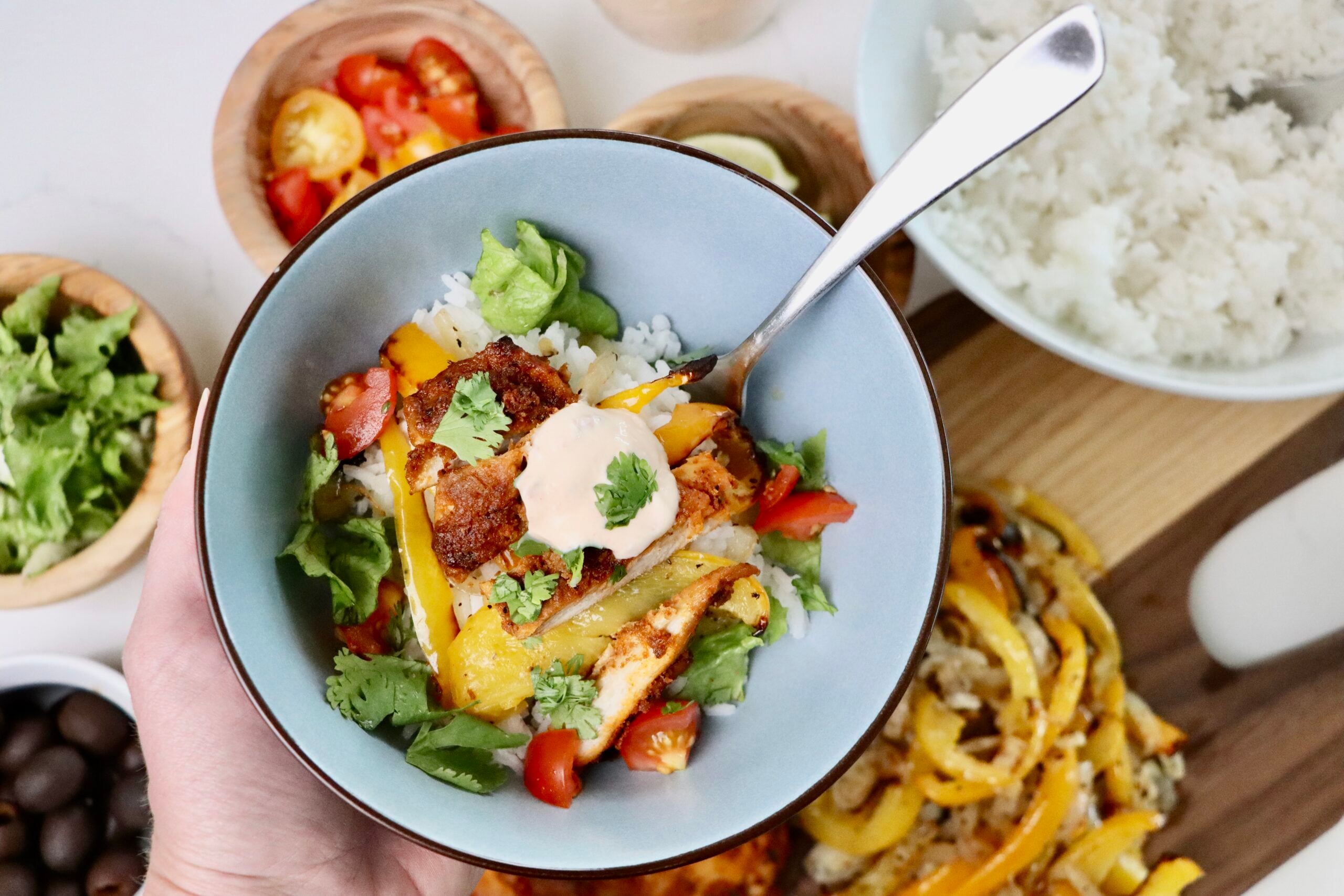 A bowl of homemade chicken fajita bowl with jasmine rice, roasted bell peppers, lettuce, tomatoes, and a dollop of chipotle crema, garnished with fresh cilantro. Surrounded by fresh toppings and ingredients on a white background.