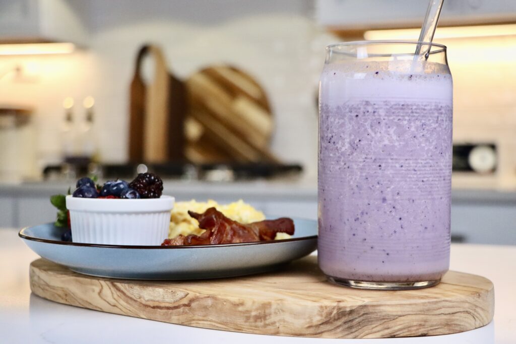 Morning Milkshake served in a glass next to a breakfast plate with scrambled eggs, crispy bacon, and fresh mixed berries on a cutting board.