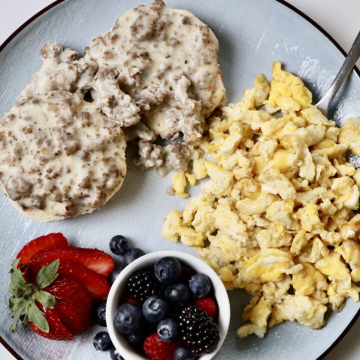 Fluffy sourdough biscuits topped with creamy homemade sausage gravy, served with scrambled eggs and fresh mixed berries on a breakfast plate.