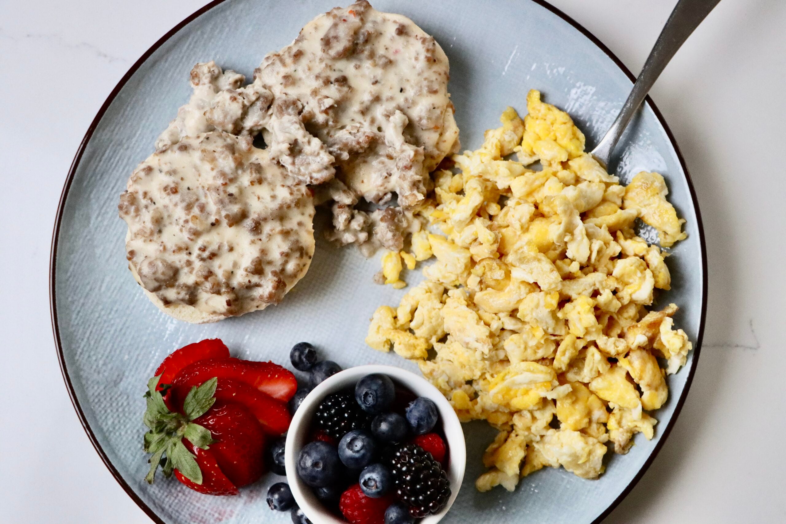 Fluffy sourdough biscuits topped with creamy homemade sausage gravy, served with scrambled eggs and fresh mixed berries on a breakfast plate.