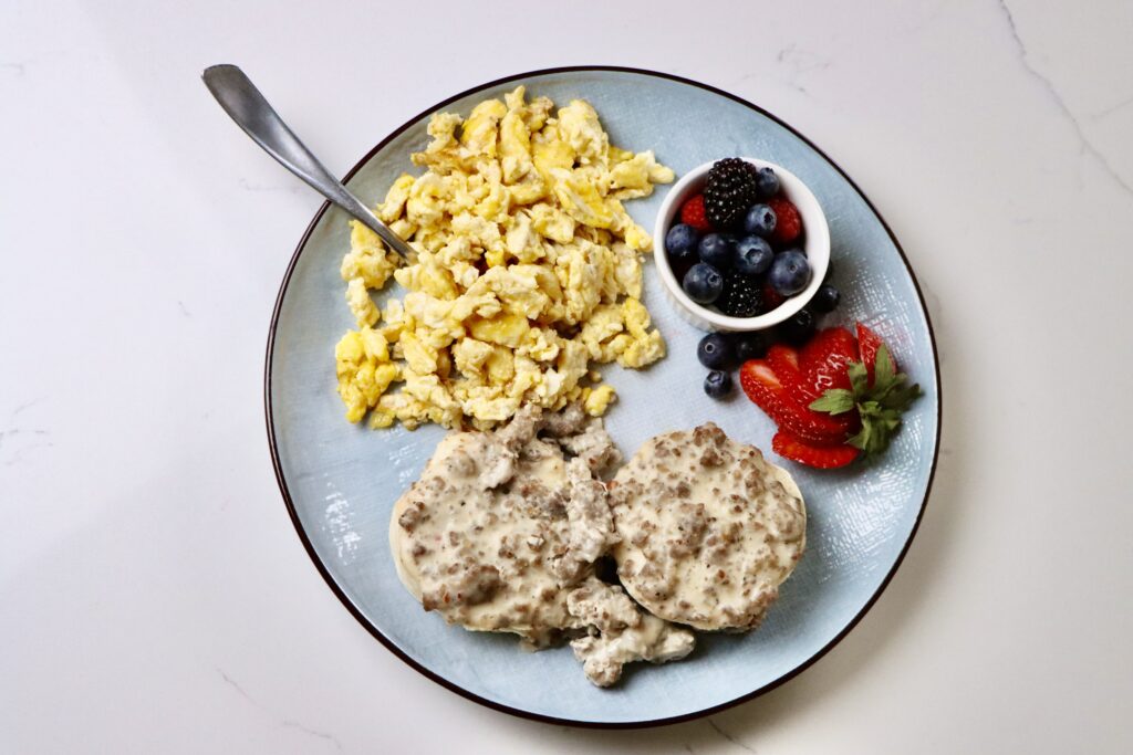 Fluffy sourdough biscuits topped with creamy homemade sausage gravy, served with scrambled eggs and fresh mixed berries on a breakfast plate.