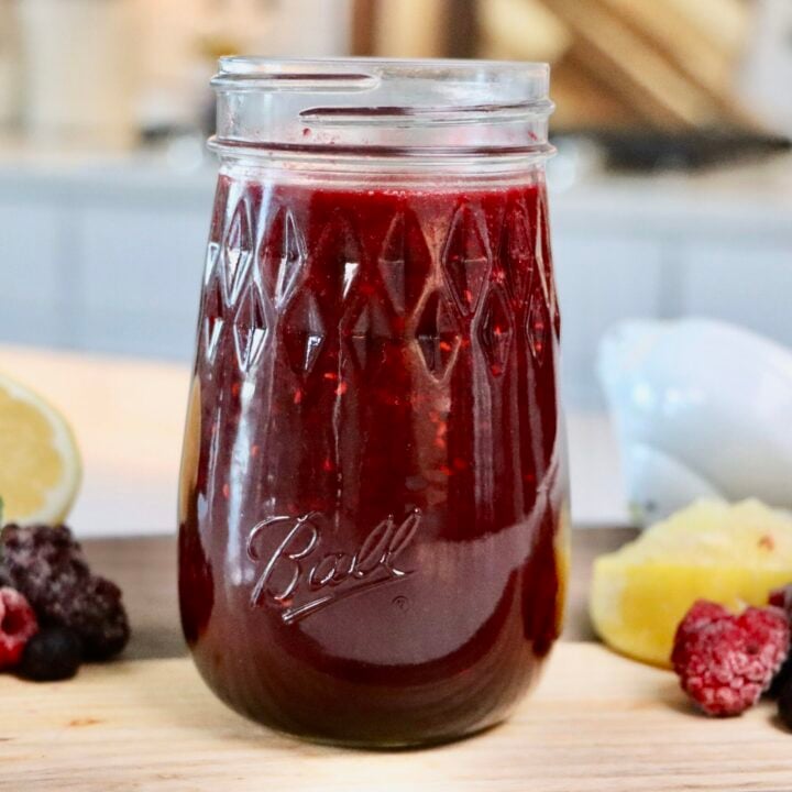 Glass jar filled with thick homemade berry sauce made from whole mixed berries, sitting on a white countertop with a spoon and fresh berries nearby.