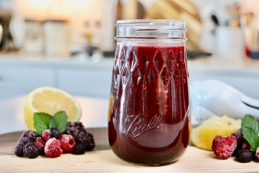 Glass jar filled with thick homemade berry sauce made from whole mixed berries, sitting on a white countertop with a spoon and fresh berries nearby.