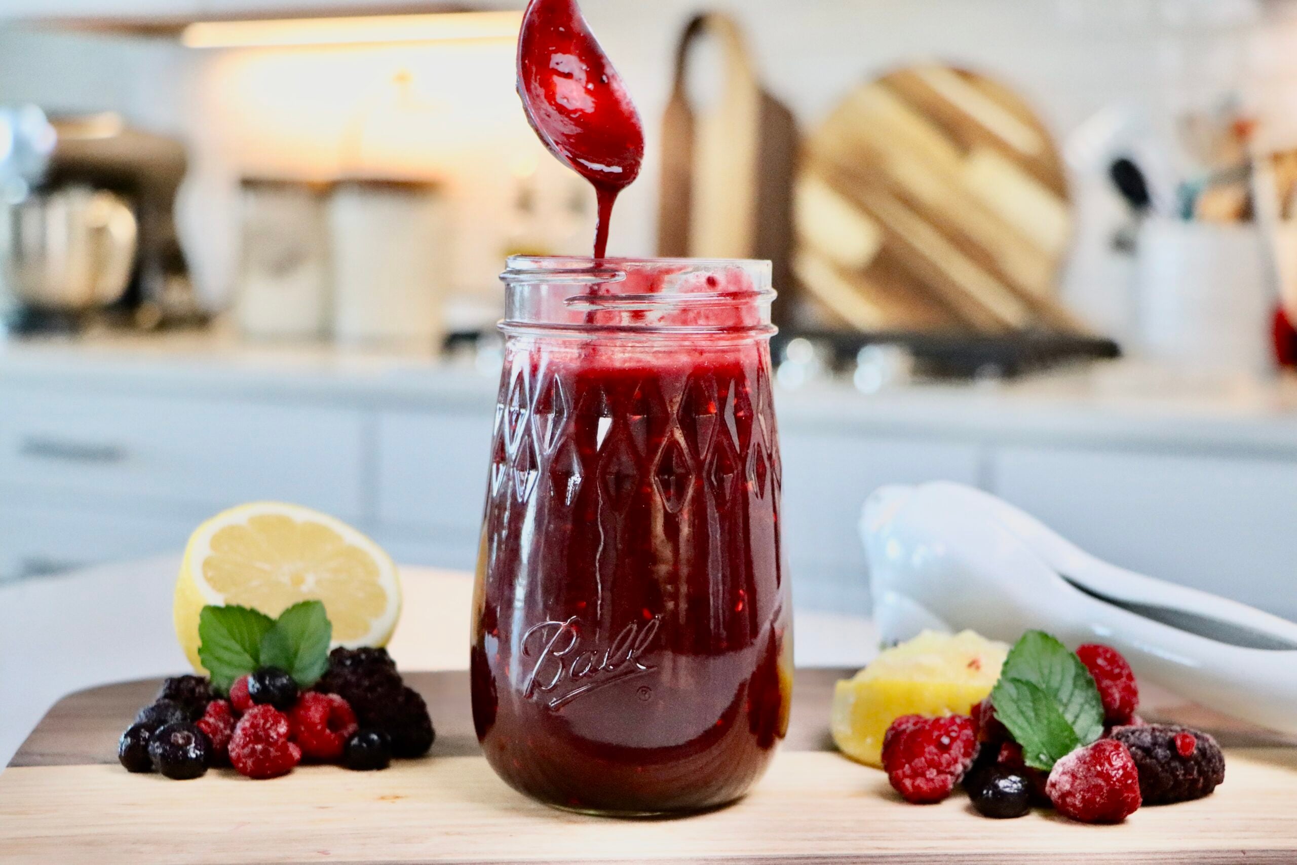 Glass jar filled with thick homemade berry sauce made from whole mixed berries, sitting on a white countertop with a spoon and fresh berries nearby.