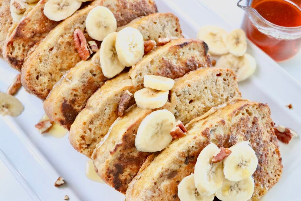 Slices of Banana Bread French Toast sitting on a white tray with sliced bananas and pecans on top.