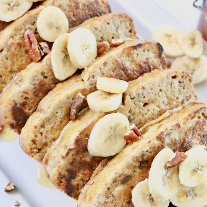 Slices of Banana Bread French Toast sitting on a white tray with sliced bananas and pecans on top.