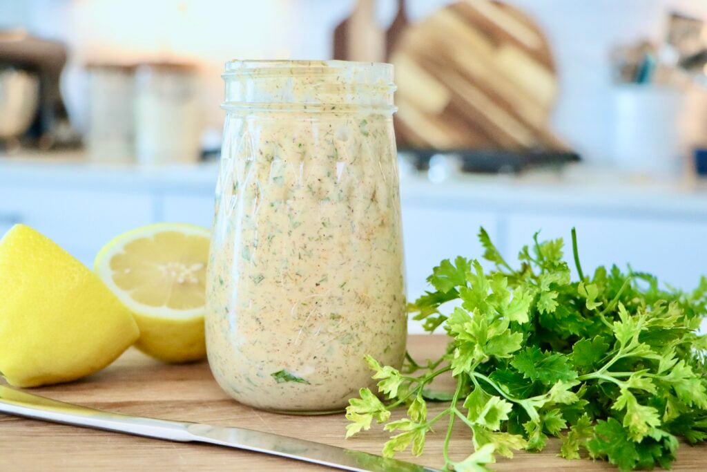 Herb Mayo in a glass jar on a cutting board next to a cut lemon and fresh parsley
