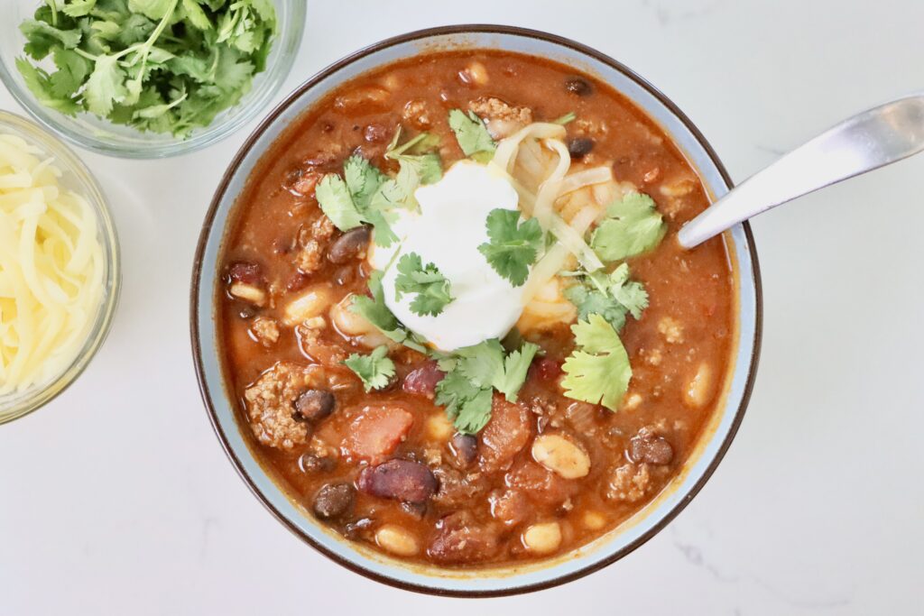 Hearty Homemade Chili in a bowl and pot next to bowls of cheese, cilantro and sour cream.