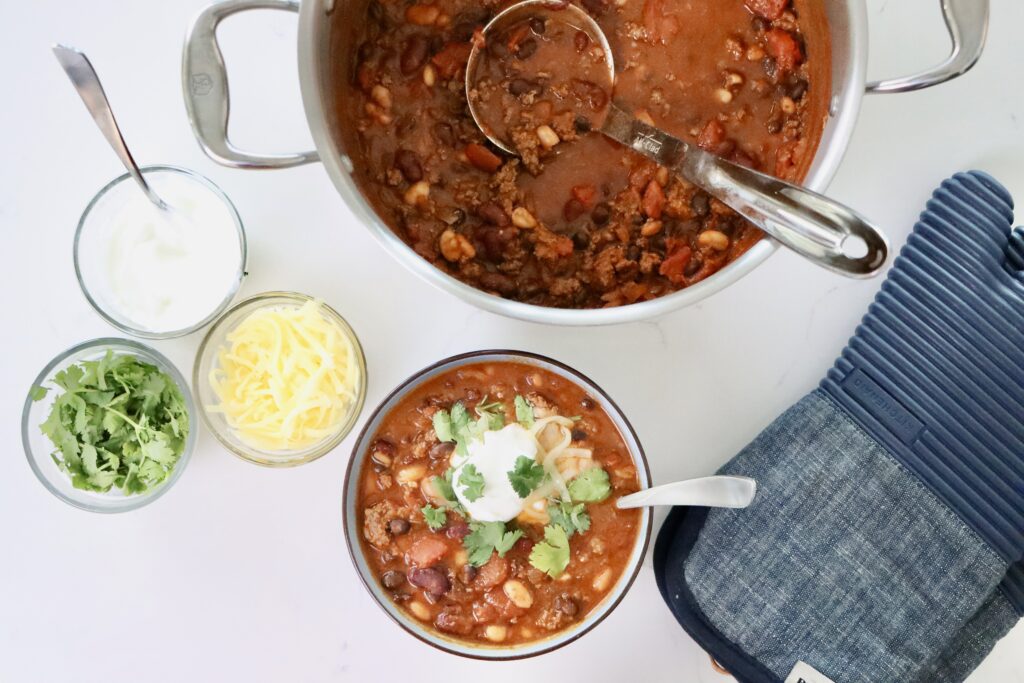 Hearty Homemade Chili in a bowl and pot next to bowls of cheese, cilantro and sour cream.