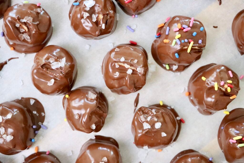 A tray of homemade no-bake peanut butter balls coated in smooth semi-sweet chocolate, some topped with colorful sprinkles and sea salt flakes, made from natural peanut butter, butter, and powdered sugar. Bite-sized, chocolatey treats made from scratch using real ingredients, perfect for holidays, gifting, or everyday snacking.