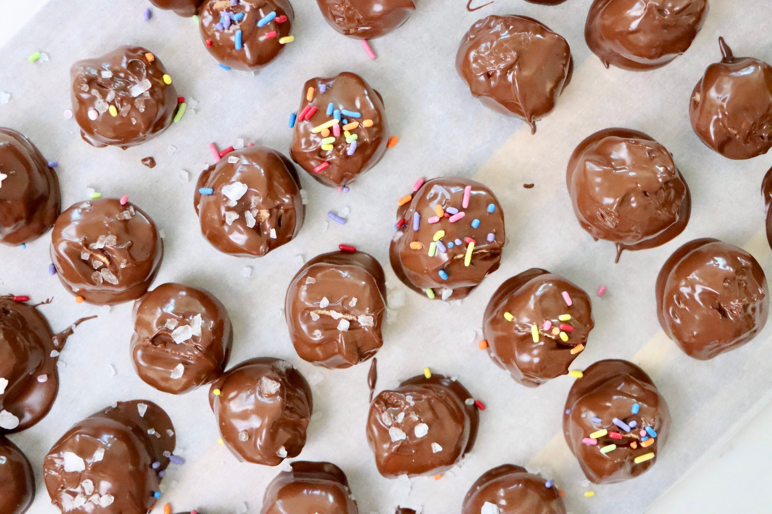 A tray of homemade no-bake peanut butter balls coated in smooth semi-sweet chocolate, some topped with colorful sprinkles and sea salt flakes, made from natural peanut butter, butter, and powdered sugar. Bite-sized, chocolatey treats made from scratch using real ingredients, perfect for holidays, gifting, or everyday snacking.