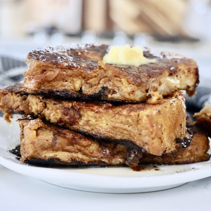 A stack of golden-brown pumpkin French toast made with thick slices of sourdough bread, topped with maple syrup, a and a sprinkle of powdered sugar.