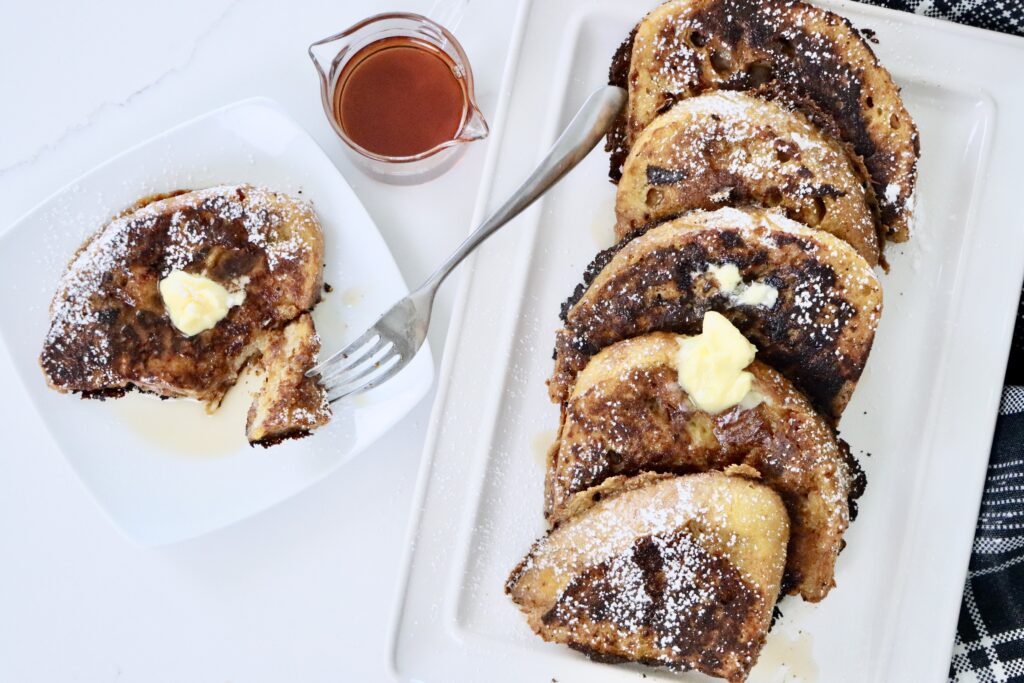 A stack of golden-brown pumpkin French toast made with thick slices of sourdough bread, topped with maple syrup, a and a sprinkle of powdered sugar.