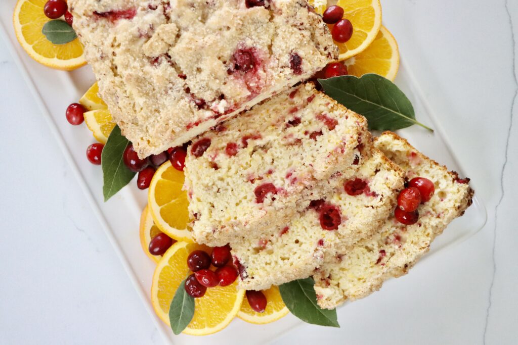 A loaf of homemade cranberry orange bread on a white tray, topped with turbinado sugar, with slices showing soft, moist texture and fresh cranberries inside.