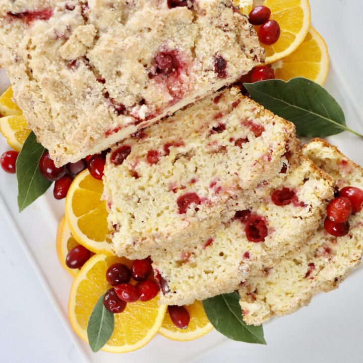 A loaf of homemade cranberry orange bread on a white tray, topped with turbinado sugar, with slices showing soft, moist texture and fresh cranberries inside.