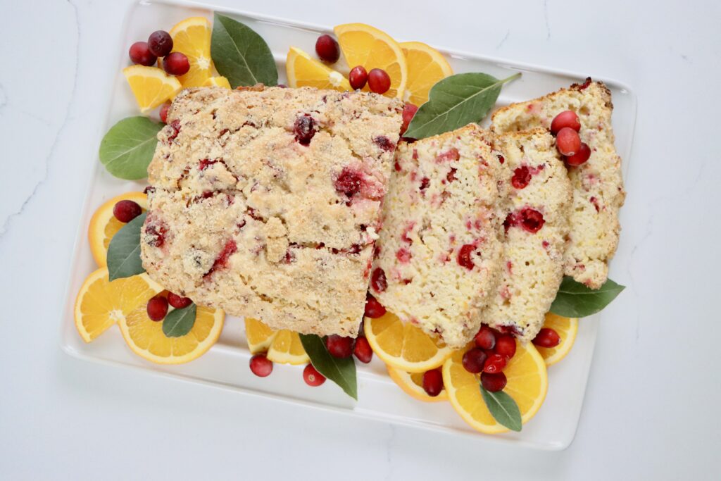 A loaf of homemade cranberry orange bread on a white tray, topped with turbinado sugar, with slices showing soft, moist texture and fresh cranberries inside.