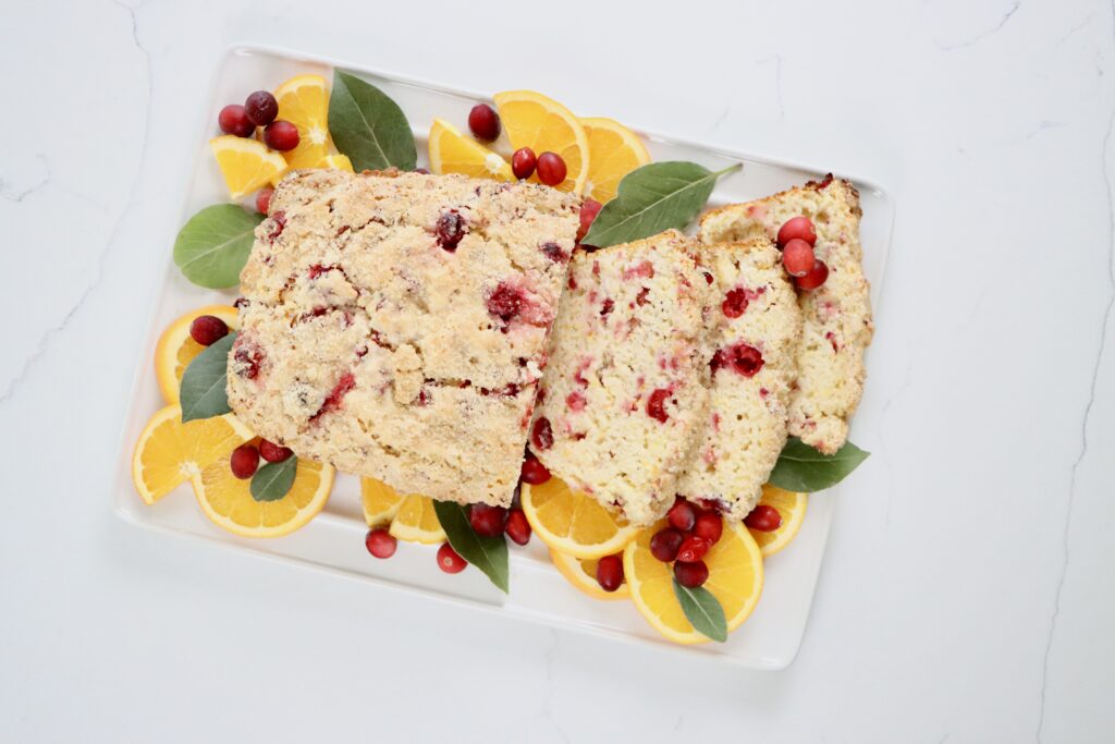 A loaf of homemade cranberry orange bread on a white tray, topped with turbinado sugar, with slices showing soft, moist texture and fresh cranberries inside.