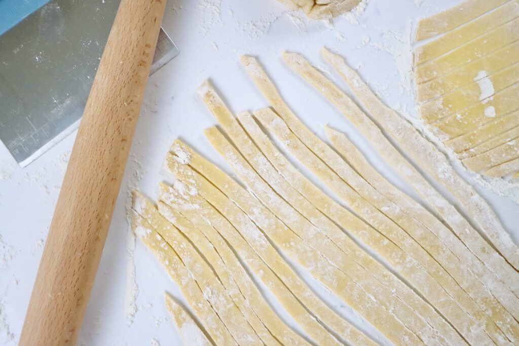 Fresh pasta being cut into fettuccine noodles by hand on a floured countertop, from scratch at home.